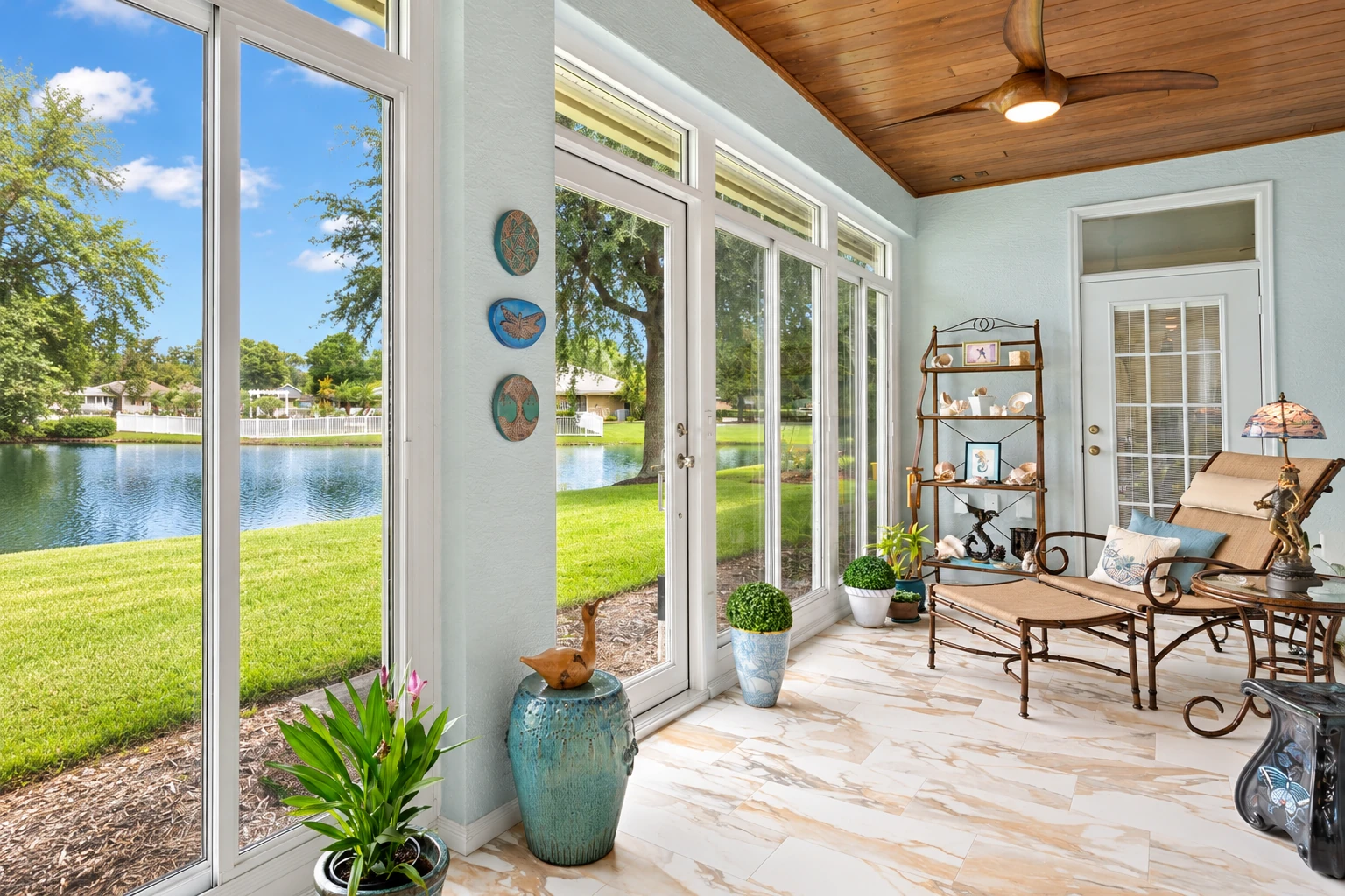 A bright four-season sunroom has white-framed windows, a wood ceiling, and seating overlooking a lake.