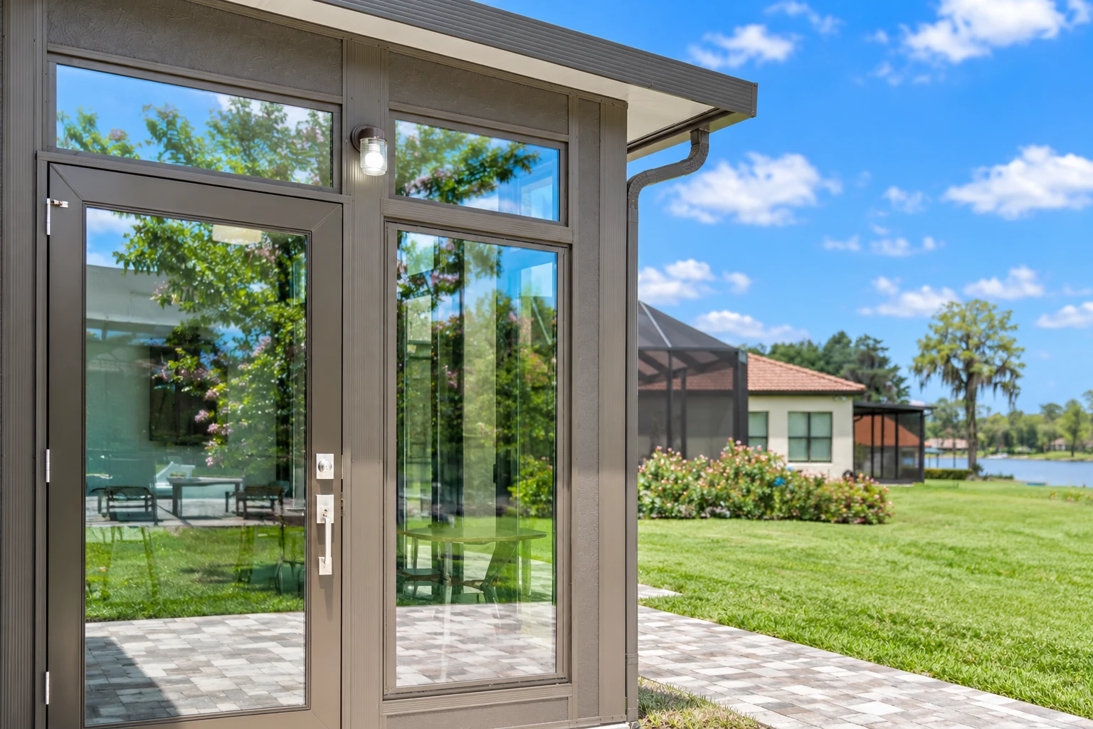 A dark-framed sunroom addition has tall glass doors and windows beside a paved patio and green lawn.