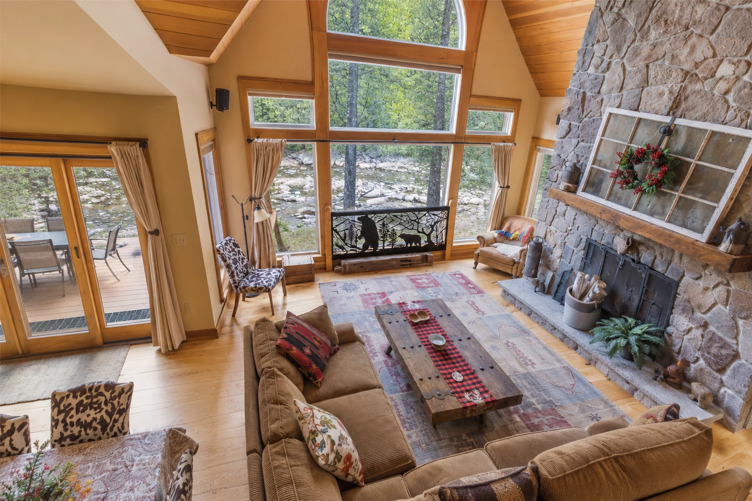 A vaulted sunroom with timber beams and tall windows features a stone fireplace, wood floors, and cozy seating around a coffee table.